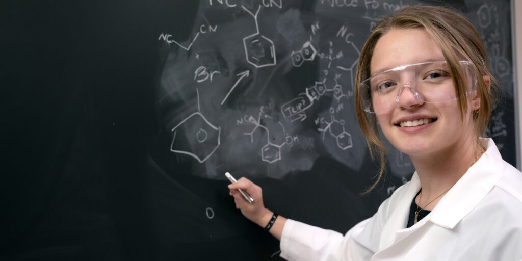 student in lab coat writing on chalkboard